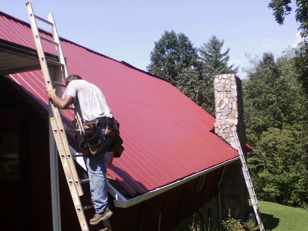 Roofer on a ladder beside a red metal roof and stone chimney — Soffit & Fascia Repair by Roofing Brothers in Boscobel